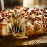 Freshly baked Cheesy Bacon Ranch Pull-Apart Bread on a rustic wooden board, topped with crispy bacon and gooey melted cheese. A piece is being pulled apart, showcasing the stretchy cheese and golden-brown crust. Warm, inviting, and perfect for appetizers or party snacks